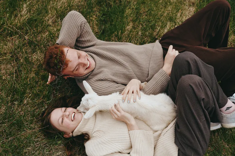 Couple outdoors, relaxed and affectionate, cozy moment on grass with a fluffy dog, warm sweaters, natural colors, gentle breeze, sunlit mood, lifestyle scene
