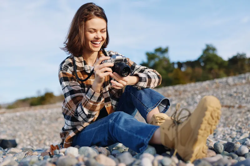 Woman photographer on a rocky beach takes a candid shot with a compact camera, wearing a plaid shirt and boots, smiling under a bright sky during outdoor photography