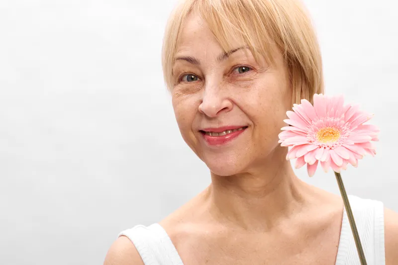 Woman smiling pink flower senior portrait, blonde hair, closeup on white studio background, gentle expression, soft lighting, calm mood, fresh beauty, minimal composition