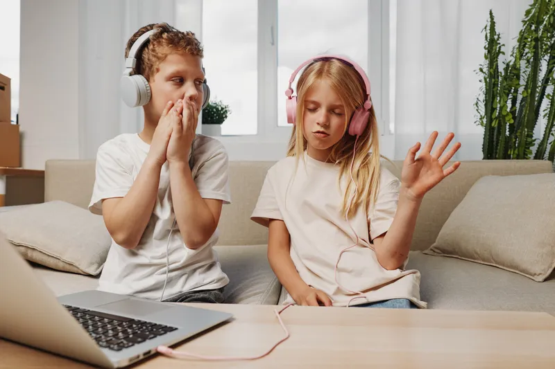 Two children wearing headphones listen to music and enjoy a relaxing moment on a cozy couch in a bright living room.