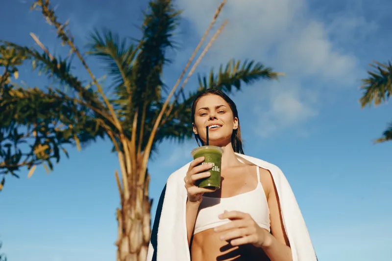 Young woman with dark hair enjoys a green smoothie outdoors under palm trees, wrapped in a white towel with a bright blue sky in the background, expressing health and relaxation.
