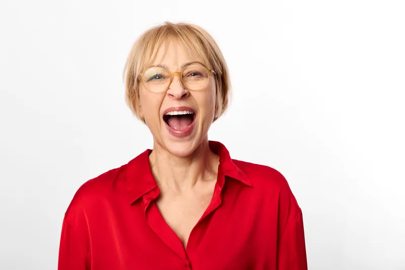 Elderly woman in red shirt and glasses, yelling with open mouth, expressing loud laughter, close up portrait against plain white background, expressive emotion