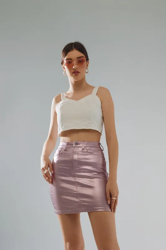 Young woman in a white crop top and metallic pink skirt poses confidently against a plain background. Fashion concept emphasizing beauty and modern style.