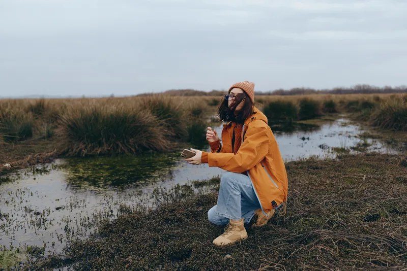 Woman lifestyle outdoors, sitting near marsh water, wearing orange jacket, scarf and boots, cloudy sky, relaxed smile, calm autumn nature, wetland landscape