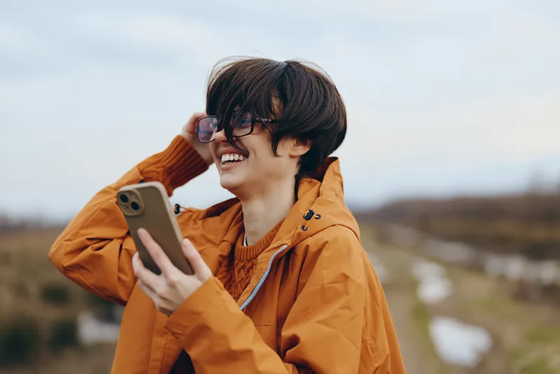 woman lifestyle outdoors smartphone coat orange smile portrait young jacket, smiling woman in orange hooded coat holds smartphone near countryside field, daylight, depth of field