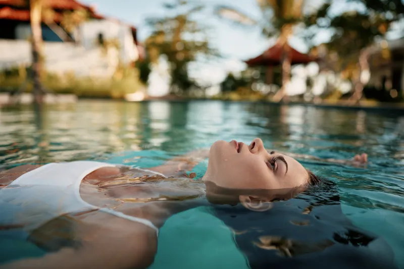 Relaxed woman floating in a pool on a sunny day, white swimsuit, tranquil turquoise water, palm trees in the background, lifestyle scene about self care, wellness, and leisure.