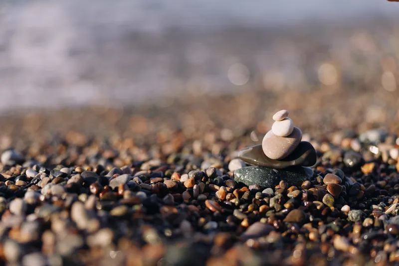 Pebbles on a sandy shore with a small stack of smooth stones forming a balanced tower, captured in soft light by the water, evoking calm and minimalist nature.