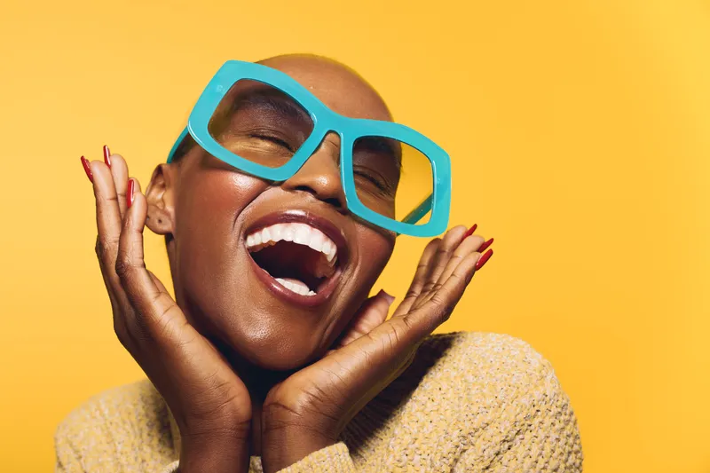 Happy dark skinned black woman with shaved head smiles widely, wearing large blue sunglasses and beige sweater. Bright yellow background creates joyful and stylish mood for fashion concept.