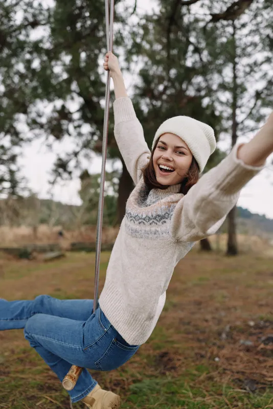 Woman on a swing outdoors wearing a cozy sweater and beanie, smiling with arms raised as she enjoys a playful moment in a park surrounded by trees and open space.