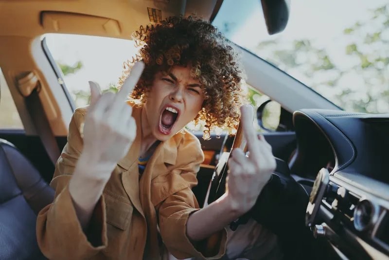A woman with curly hair sits behind the wheel, expressing bold attitude in a retro film color mood. A candid road trip concept highlighting empowerment and rebellious energy.