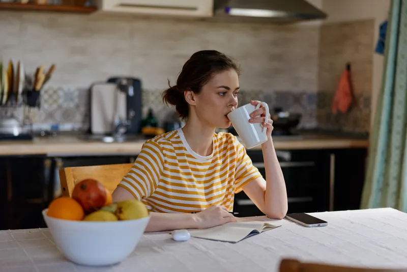 Woman enjoying a morning beverage at a home kitchen, striped shirt, soft light, fruit bowl nearby, relaxed and focused, everyday life captured in a calm cozy setting