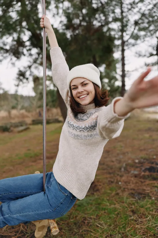 Woman on a swing in an outdoor setting, wearing a knit sweater and beanie, smiling and reaching forward, capturing a carefree playground moment amid trees and open space.