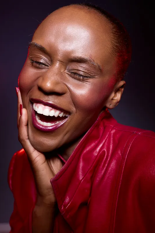 Close-up of a happy black woman with dark skin laughing joyfully, wearing a red leather jacket against a dark background. Concept of happiness, beauty, and fashion expression.