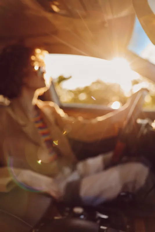 Woman driver sits in a car at sunset, embracing retro film color with warm interior glow, candid travel lifestyle moment. Natural bright sun, warm metallic highlights, relaxed posture.