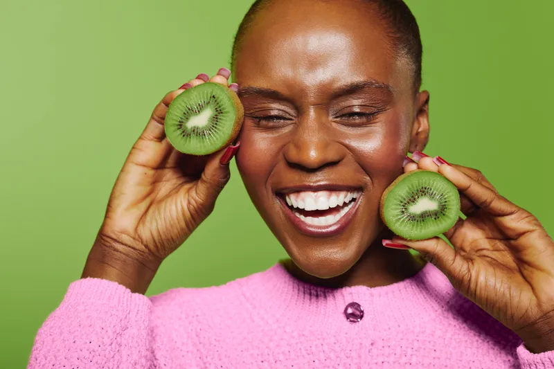 Close-up of happy African woman with dark skin holding fresh kiwi slices near face. Smiling female model in pink sweater on green background. Concept of healthy lifestyle and natural food.