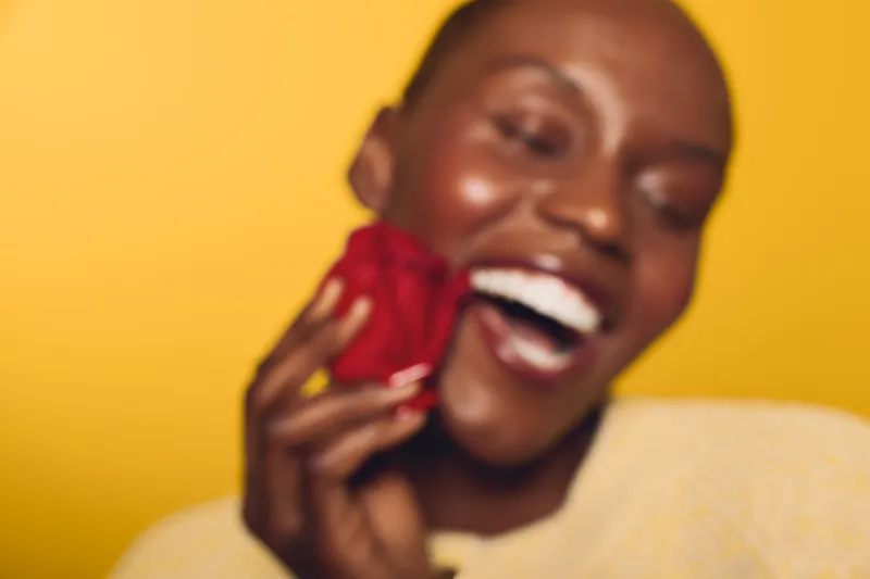 Close-up of smiling black woman with dark skin holding red rose near face on yellow background. Concept of happiness, beauty, and natural skincare with vibrant colors and joyful mood.