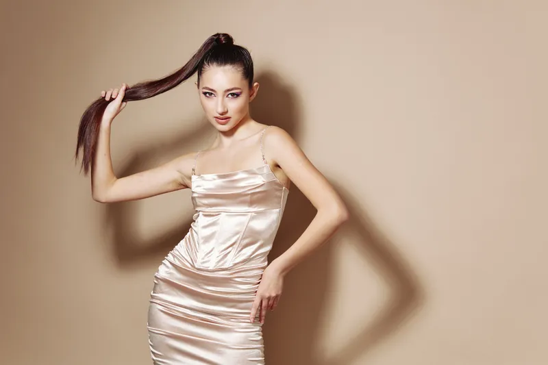 Young woman in a shiny satin dress holds her long ponytail while posing confidently against a beige background. Concept for beauty, fashion, and creative style portrait.
