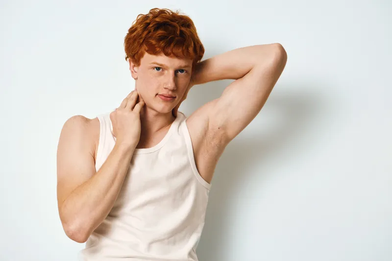 Young man with red hair posing in tank top against white background, showing casual style and confident expression with hand touching neck and arm raised.