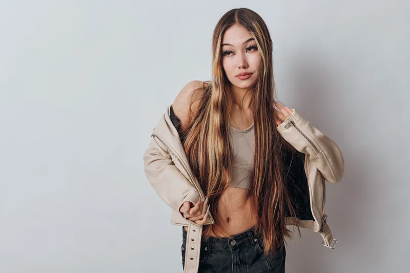 Young woman posing with long hair and casual jacket on gray background, wearing black jeans and crop top. Fashion portrait with confident expression in studio lighting.