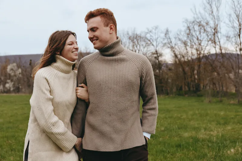 Young couple in cozy knit sweaters walks arm in arm through a grassy field during autumn, sharing a warm moment and smiling at each other with trees in the background.