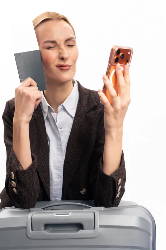 Young woman with suitcase holding passport and smartphone, making a funny face. Travel concept showing excitement and preparation for trip, isolated on white background.