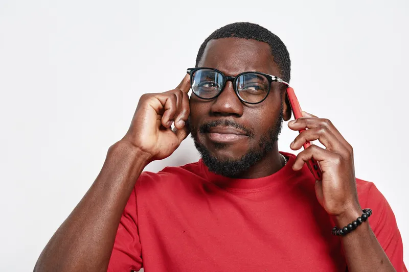 African American man with dark skin wearing glasses and red shirt talking on smartphone and touching temple for thinking or concentration on white background.