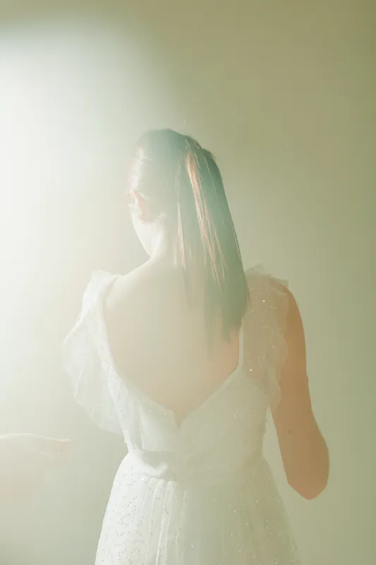 Young woman in a white dress with open back stands in soft light. Concept of beauty, fashion, and elegance with a delicate atmosphere and subtle glow.