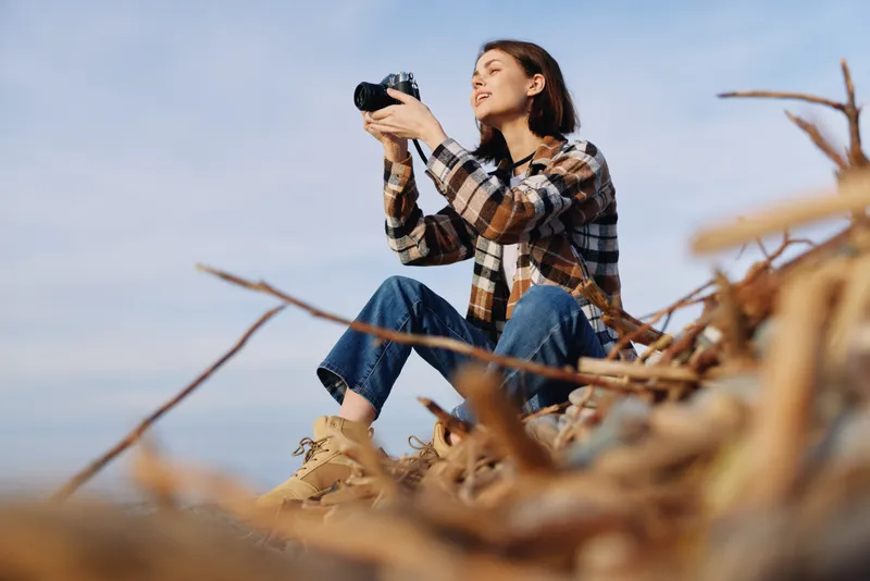 A woman photographer sits among dried grass and captures the landscape with a camera wearing a plaid shirt and jeans showcasing outdoor creativity and nature photography moments