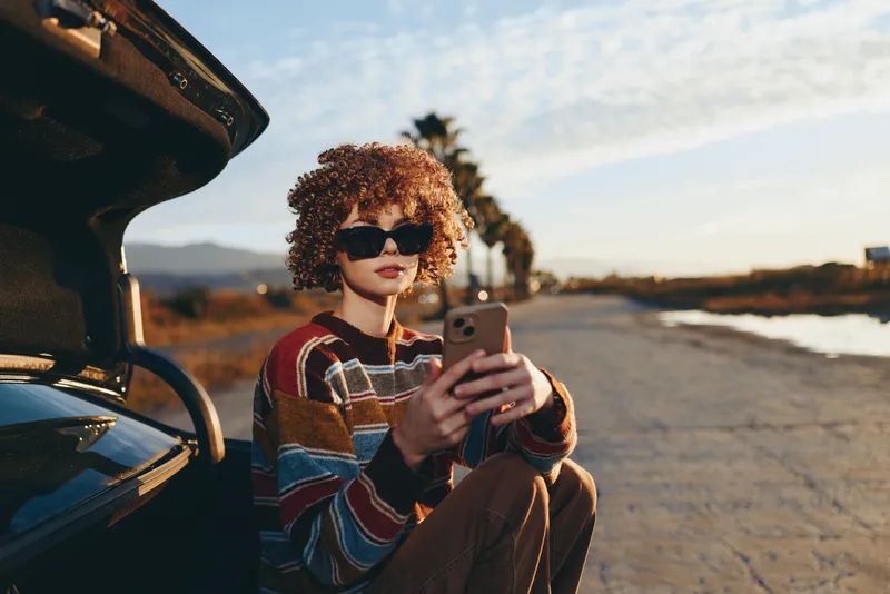 Curly haired woman wearing sunglasses taking selfie with smartphone near car on a seaside road at sunset, outdoor lifestyle concept, relaxed travel mood, soft natural light today