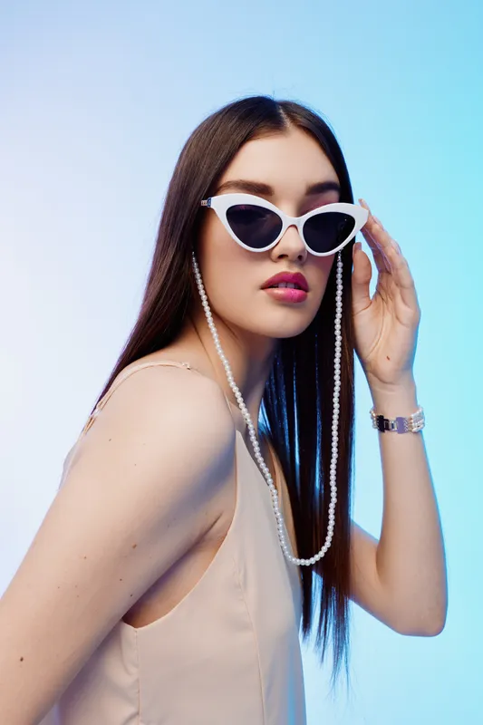 Young woman beauty fashion wearing white sunglasses and pearl necklace poses confidently against a blue background. Her long hair and stylish look create a modern vibe.