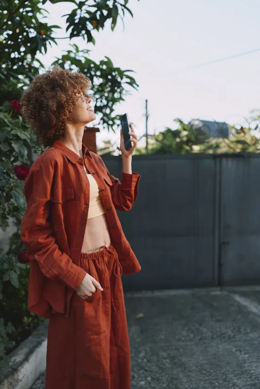 Young woman with curly hair wearing a rust suit stands outdoors among greenery, looking upward and holding a cigarette, a candid lifestyle moment with soft natural light, warm tones