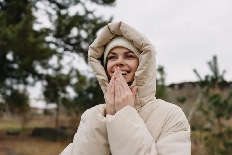 Woman in a warm hooded coat smiles with hands clasped in front of her outdoors, cold weather scene with soft natural light and trees in the background.