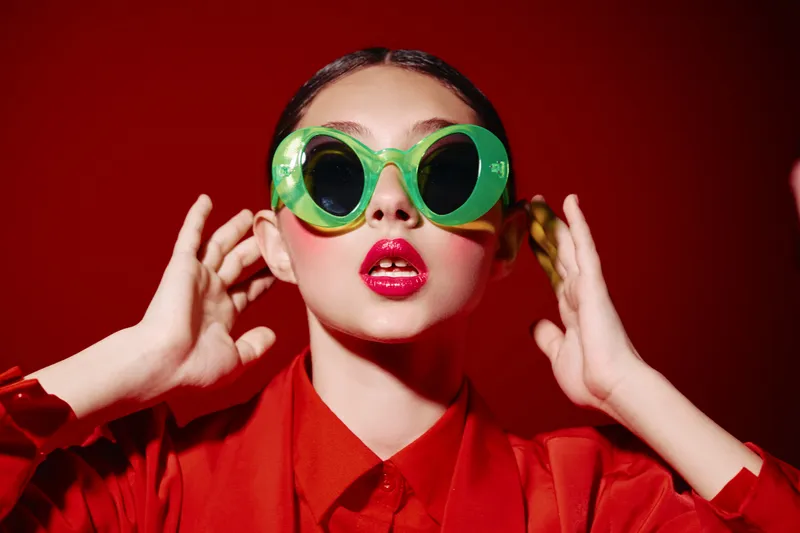 Young woman with bright makeup wearing large green sunglasses posing on red background. Creative beauty and fashion concept emphasizing bold style and color contrast.