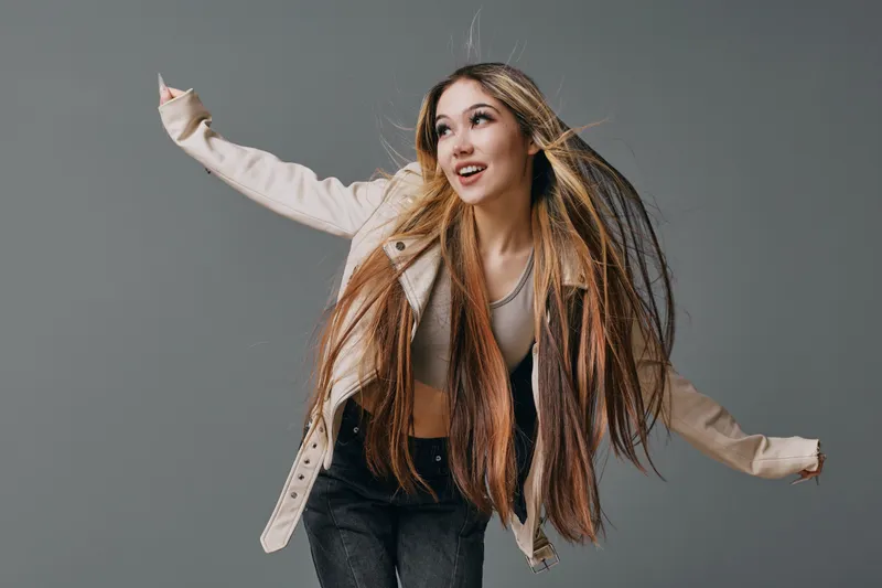 Young woman with long hair wearing beige jacket and tank top posing energetically against gray background for fashion portrait and lifestyle photography session.