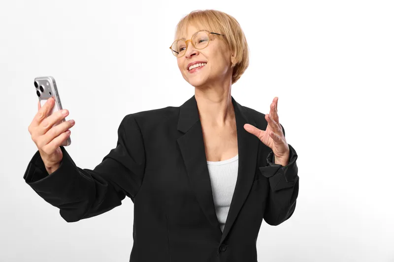 Smiling senior businesswoman holding smartphone in studio, open hand gesture, confident communication, modern technology concept, isolated on white background