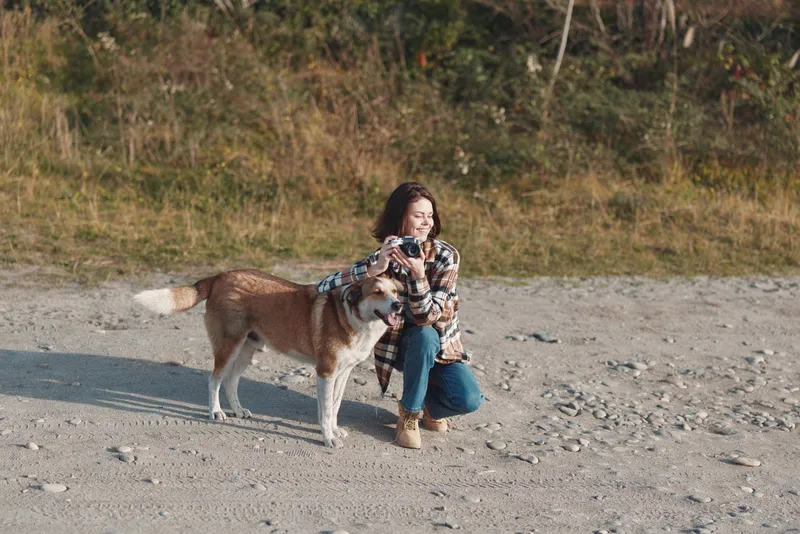 Woman kneels on a gravel beach with a large dog, petting the dog and enjoying an outdoor moment, autumn light, trees in the background, candid portrait of companionship.