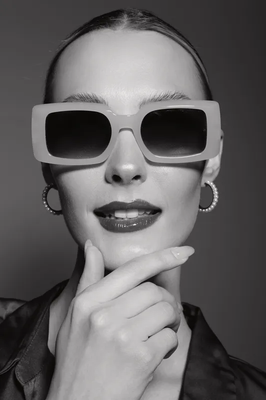 Stylish young white woman with light skin wearing large sunglasses and hoop earrings poses confidently with hand near chin in studio portrait.