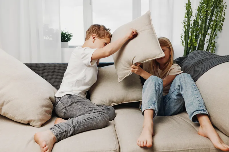Two children having fun pillow fighting on sofa in bright living room. Casual clothes and playful interaction create joyful family home moment.