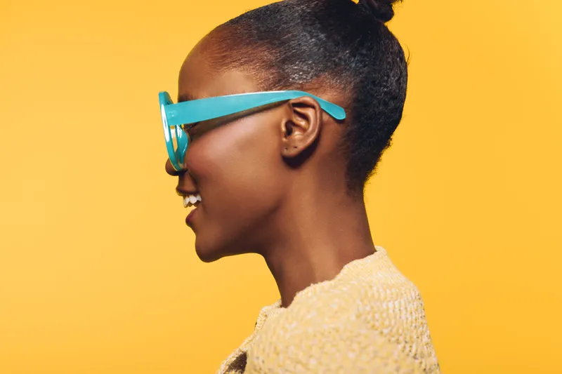 Profile of a smiling African woman with dark skin wearing blue glasses and a beige sweater. The bright yellow background creates a cheerful and vibrant portrait concept of happiness and style.