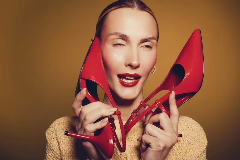 Woman with slicked hair holds red high heels near face with eyes closed and glossy lips. Studio portrait on yellow background. Concept of fashion, style, and elegance.