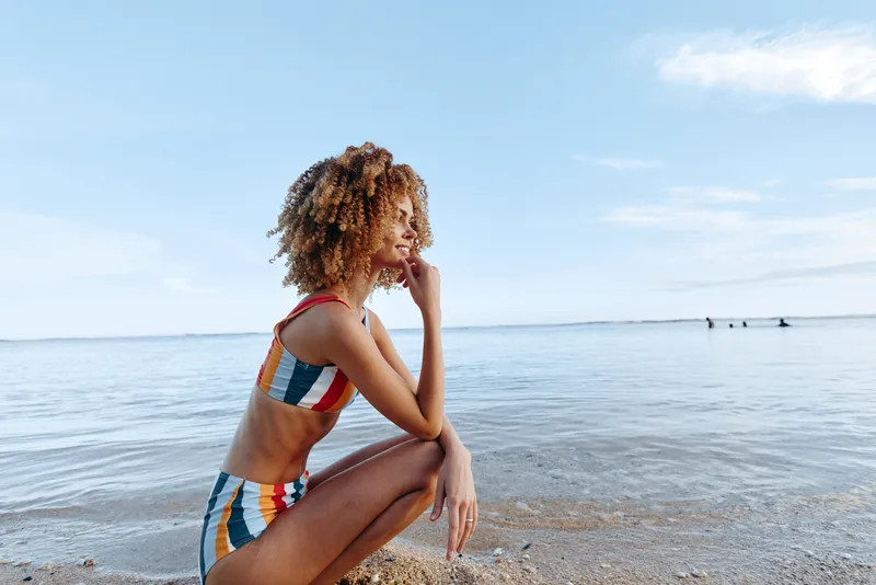 Woman sitting on beach near ocean in colorful swimsuit, head on hand, relaxing and thinking in calm summer light, gentle waves and bright sky, vacation lifestyle sea breeze peace today