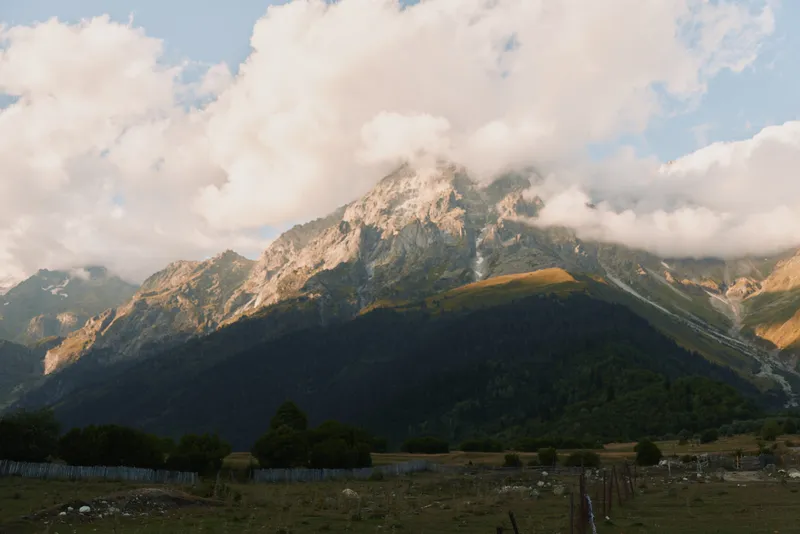 Mountain landscape with rugged peaks, scattered clouds, sunlit summit, green valley foreground, peaceful outdoor scene, natural colors, wide scenery