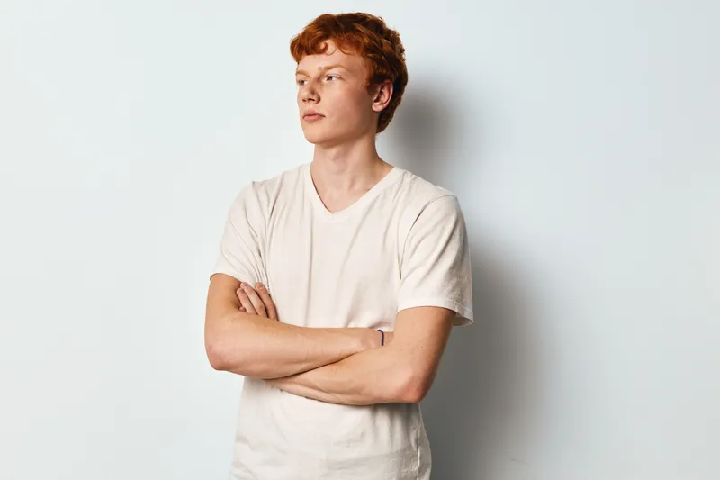 Young man with red hair and folded arms looking away in casual white t shirt against plain white background. Profile portrait of confident male model with freckles.
