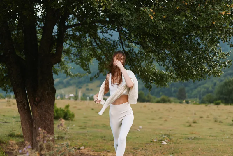 A young woman in white activewear runs joyfully under a large tree in a green field during daytime. The scene captures a carefree lifestyle and outdoor activity concept.