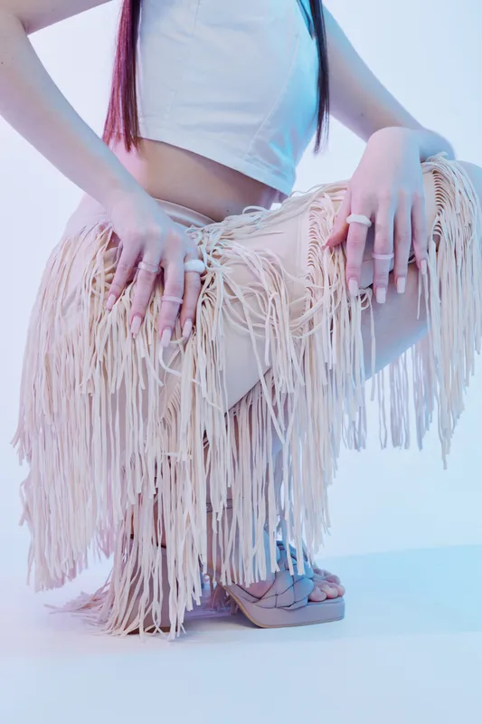 Woman in fashion outfit with fringe skirt and rings poses in studio. Concept of beauty, style, and modern trends with soft lighting and pastel tones.