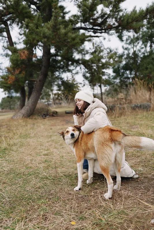 Woman in a light winter coat and white beanie crouches beside a friendly dog in a park, with autumn grass and pine trees in the background, capturing a calm moment of companionship.