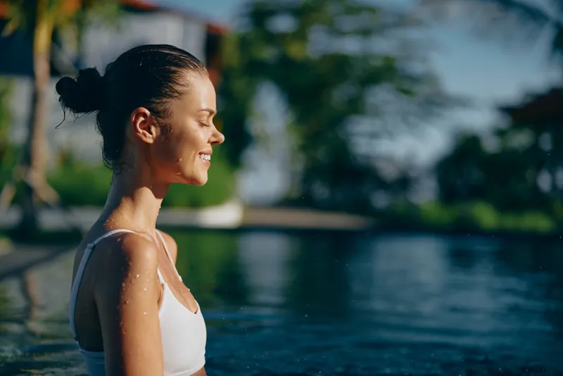 Cheerful woman by a sunlit pool, enjoying outdoor lifestyle, relaxed posture, warm breeze, bright water, lush greenery, natural colors, and a peaceful summer vibe with airy mood