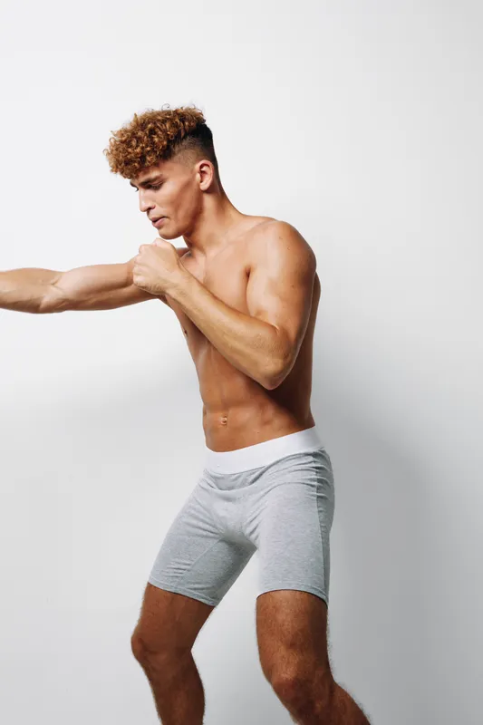 Athletic young man with curly hair practicing boxing punch in gray shorts on white background. Fitness, training, muscle, workout and sport concept with focused expression.