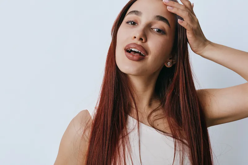 Young woman with long red hair posing with hand on head and open mouth against white background. Portrait of female model with natural makeup and casual sleeveless top.