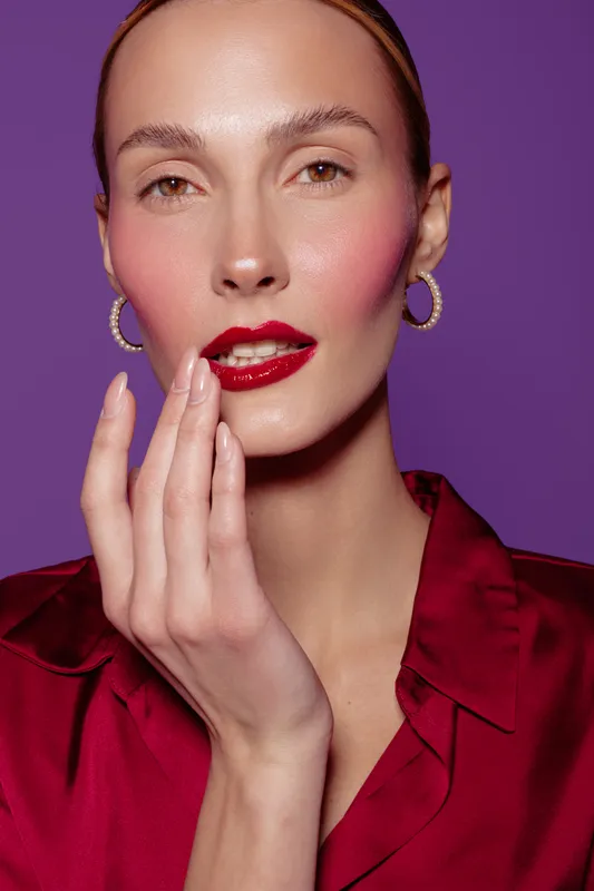 Close-up portrait of a woman with red lipstick and pink blush wearing gold hoop earrings and a red satin blouse. She touches her face gently against a purple background. Beauty and fashion concept.